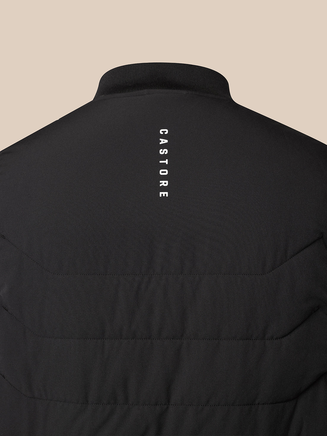 Close-up back view of a black quilted jacket with a vertical white 'CASTORE' logo printed on the upper back, set against a solid beige background.