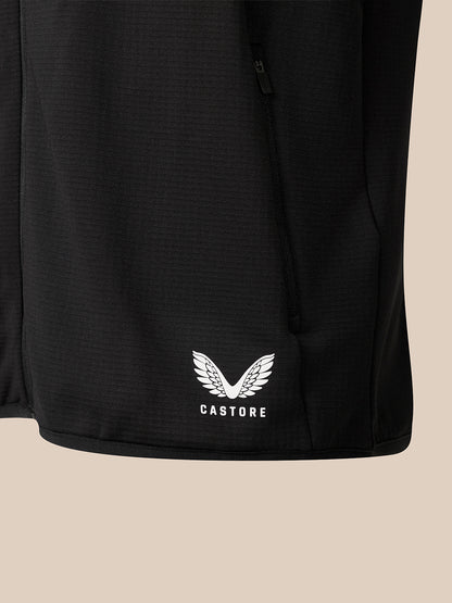Close-up of a black athletic jacket or vest with a white winged logo and the word CASTORE printed near the lower left front, shown against a plain warm beige background.