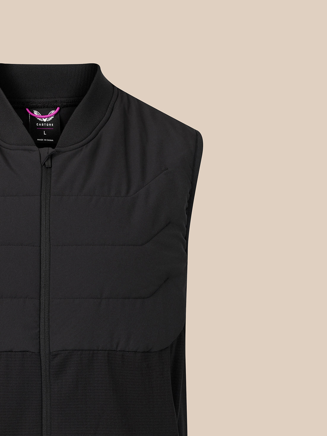 Close-up of a black sleeveless zip-front vest with horizontal quilting, shown against a plain warm beige background. A small purple and white inner neck label is visible.