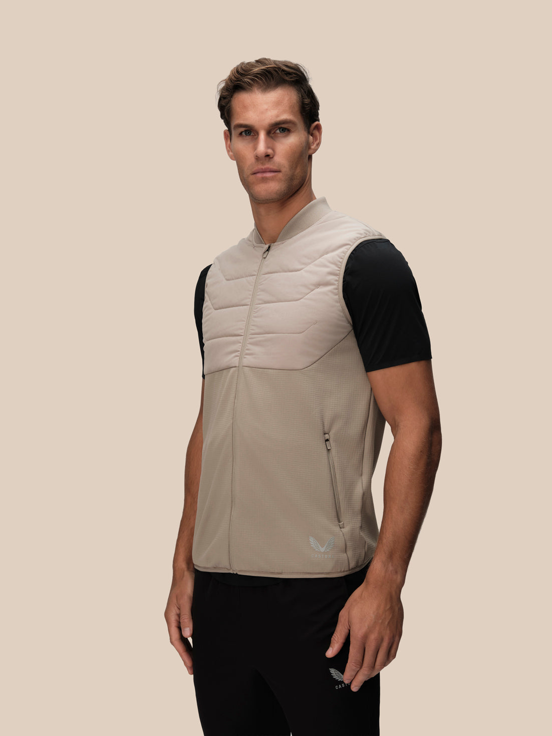 A man wearing a light beige and black color-blocked puffer vest over a black short-sleeve shirt, standing against a solid beige background.