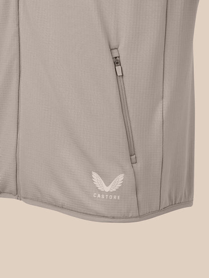 Close-up of a light gray Castore lightweight jacket with a vertical zippered pocket and the white wing logo on the lower front, set against a solid beige background.