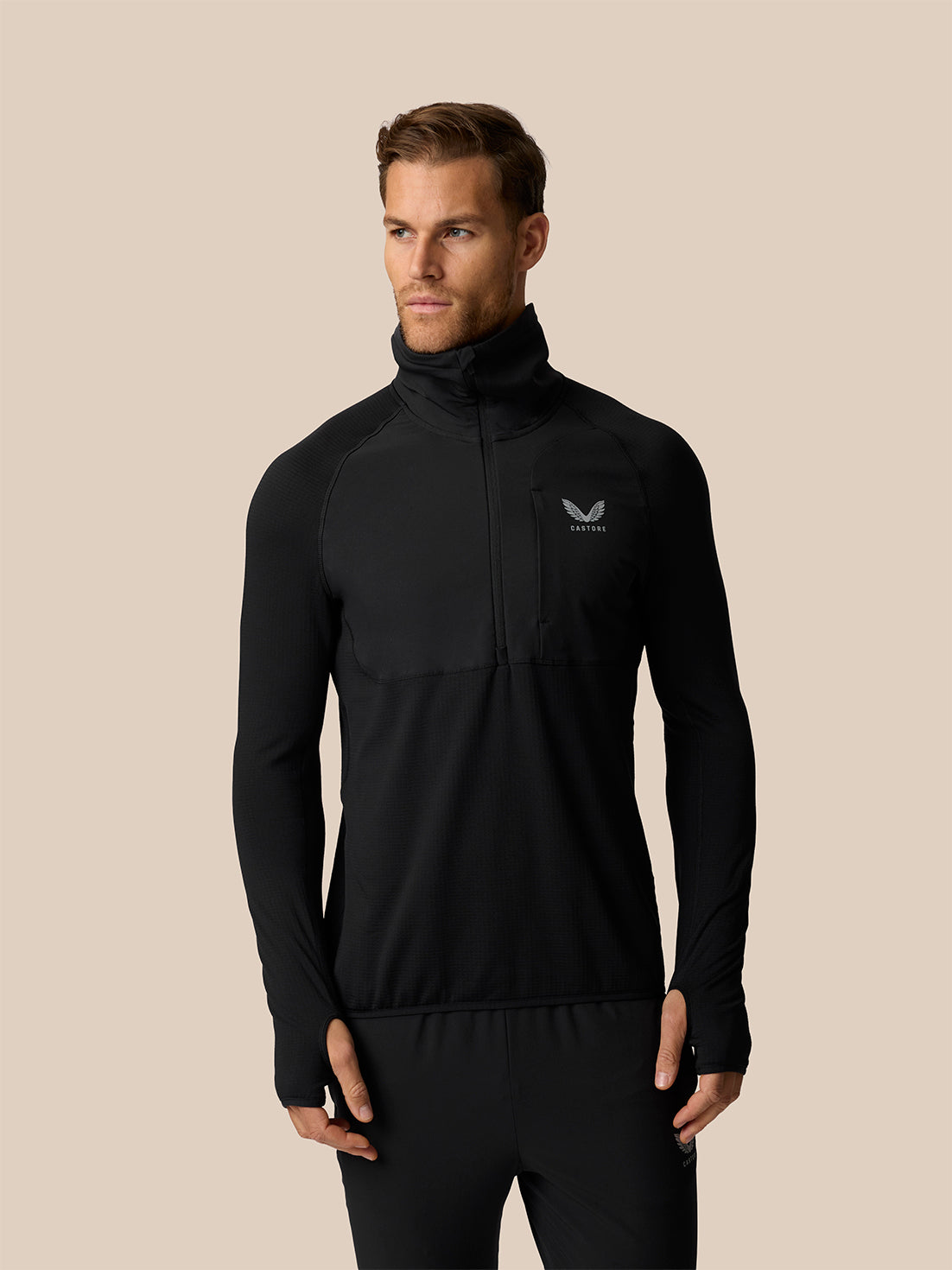 A man wearing a black long-sleeve athletic half-zip pullover with a high collar and a small white wing logo on the left chest, posed against a solid beige background.