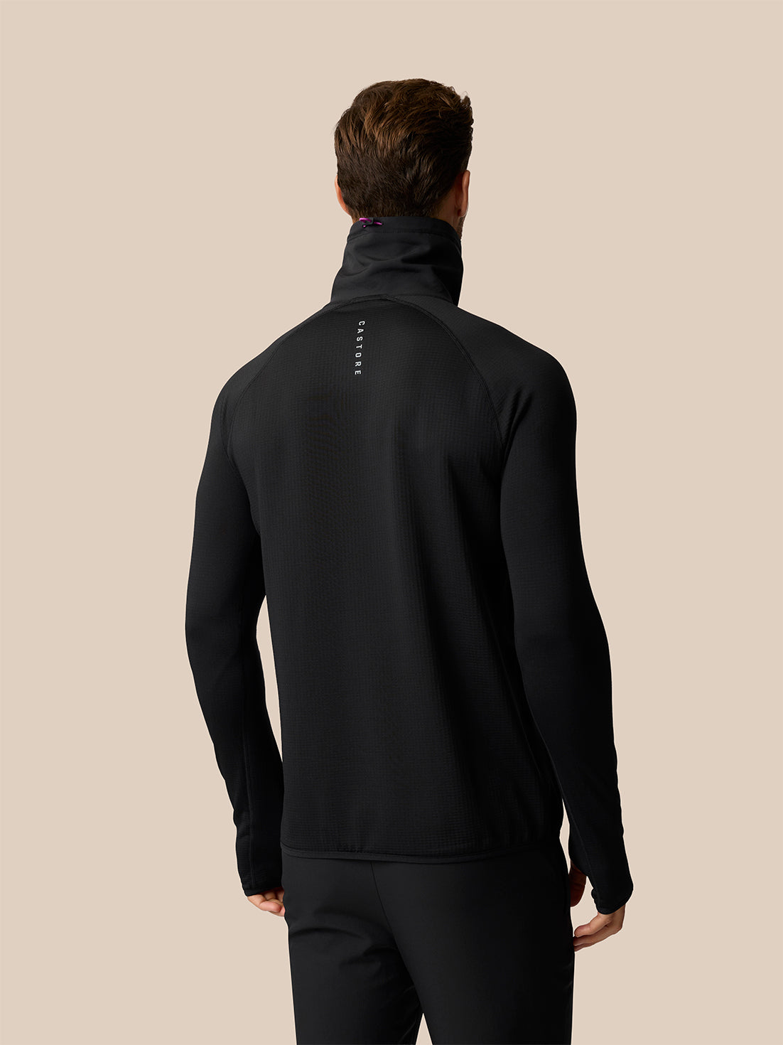 A man viewed from the back wearing a black long-sleeve athletic quarter-zip pullover with a high mock neck collar and a small vertical white logo on the upper back, set against a solid beige background.