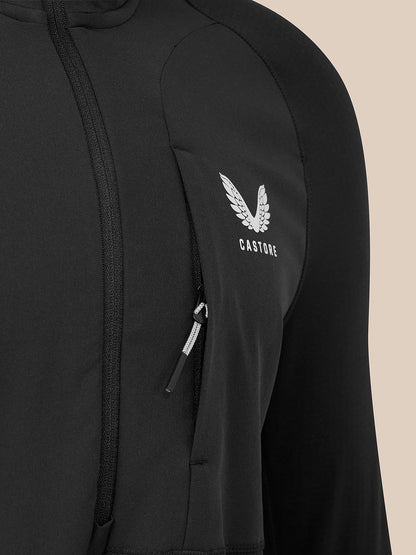 Close-up of a black Castore zip-up jacket featuring a white embroidered winged logo and brand name on the chest pocket, set against a solid beige background.