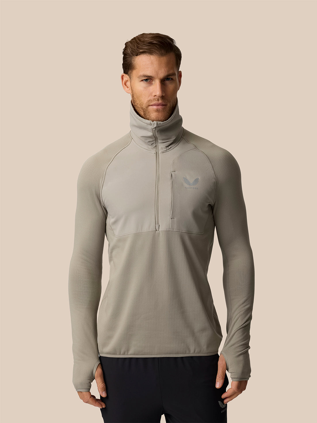 A man wearing a light gray long-sleeve quarter-zip athletic pullover with a high mock neck collar, shown against a solid beige background.