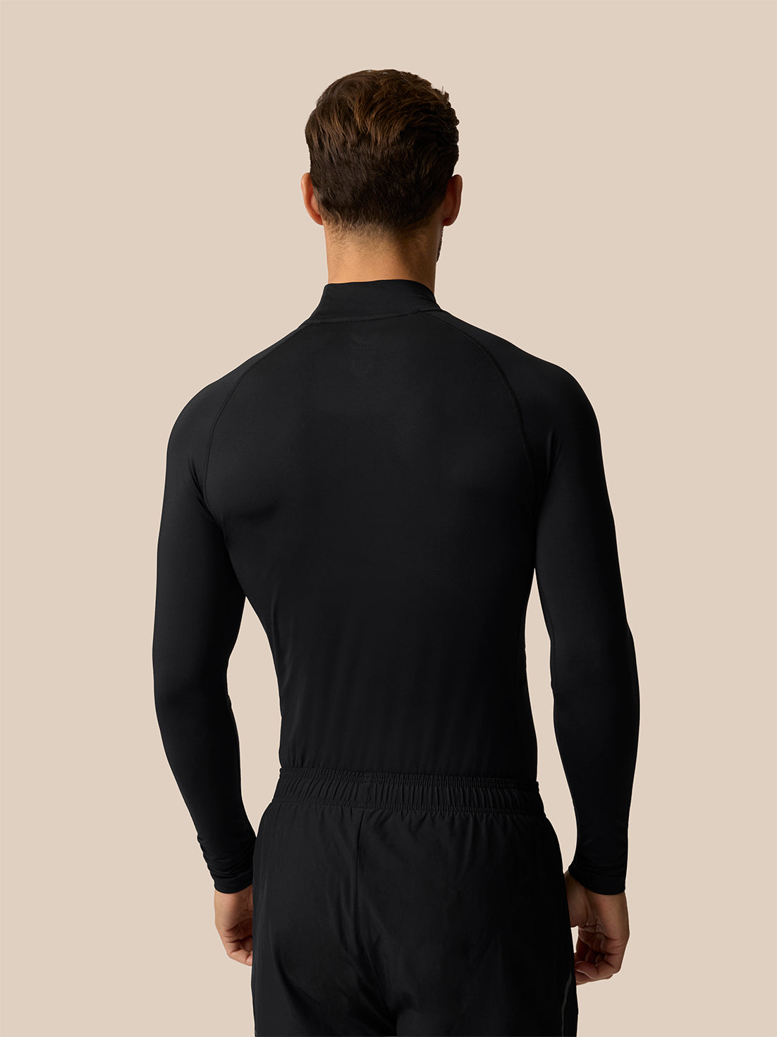 A man viewed from the back wearing a black long-sleeve mock neck athletic top and black shorts against a solid beige background.