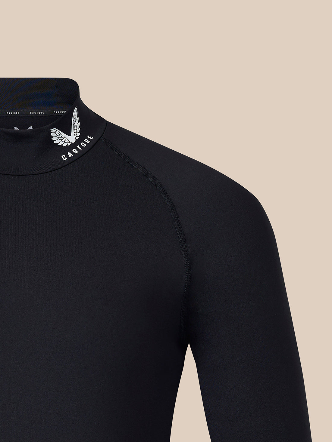 Close-up of a black long-sleeve high-neck top with a white winged logo and the word 'CASTORE' on the inner neck collar, shown against a plain beige background.