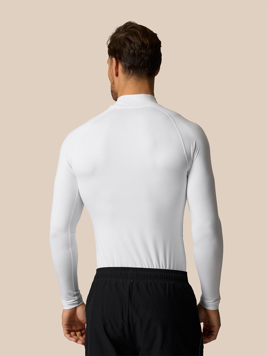 A man viewed from the back wearing a fitted white long-sleeve athletic top and black shorts against a plain beige studio background.