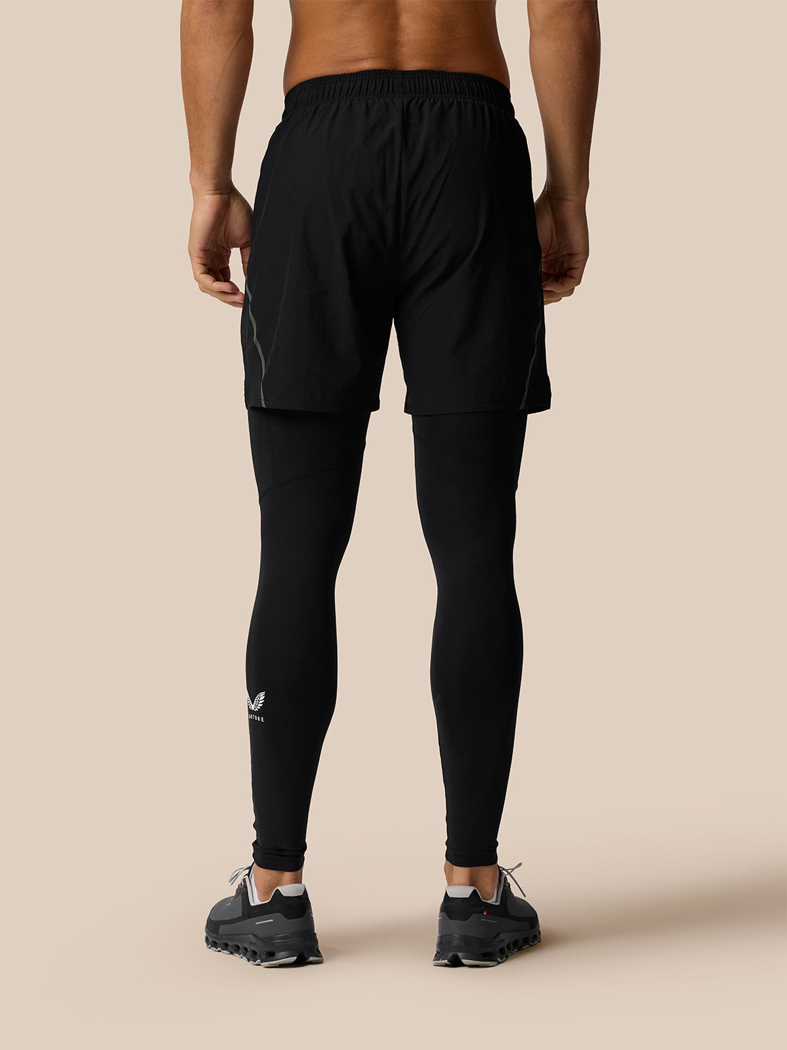 Rear view of a male model wearing black athletic shorts over a matching black full-length compression tights set, paired with black running shoes, against a solid beige background.