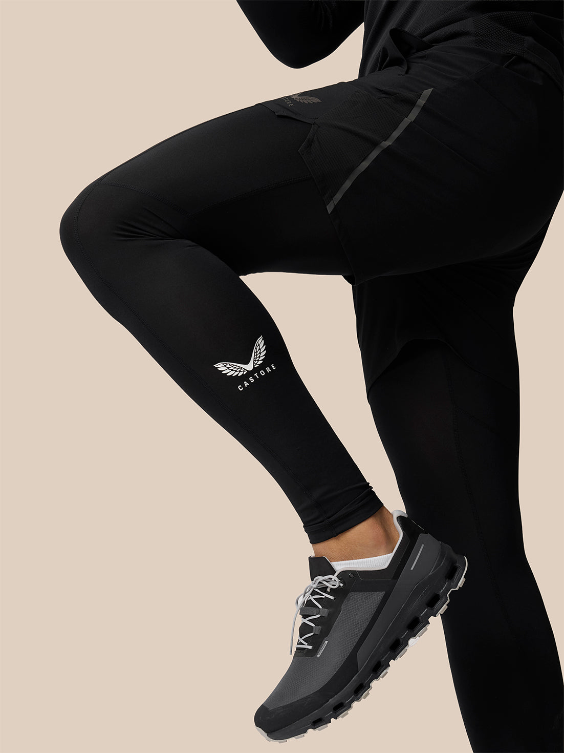 Black athletic leggings worn by a model, featuring a white wing logo and the brand name 'CASTORE' on the lower left leg, paired with dark gray trail running shoes against a solid beige background.
