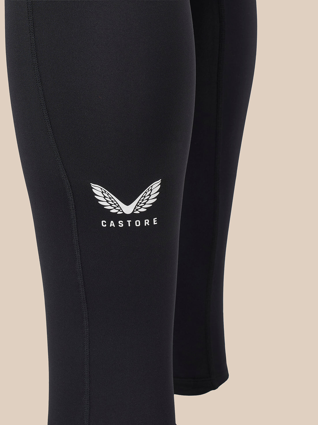 Close-up of black leggings with a white Castore wing logo and brand name printed on the upper thigh, set against a solid beige background.