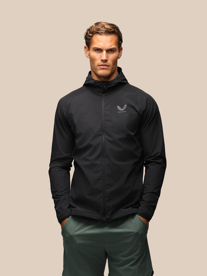 A man wearing a black hooded zip-up jacket with a small gray wing logo on the left chest, paired with dark green pants, standing against a solid beige background.