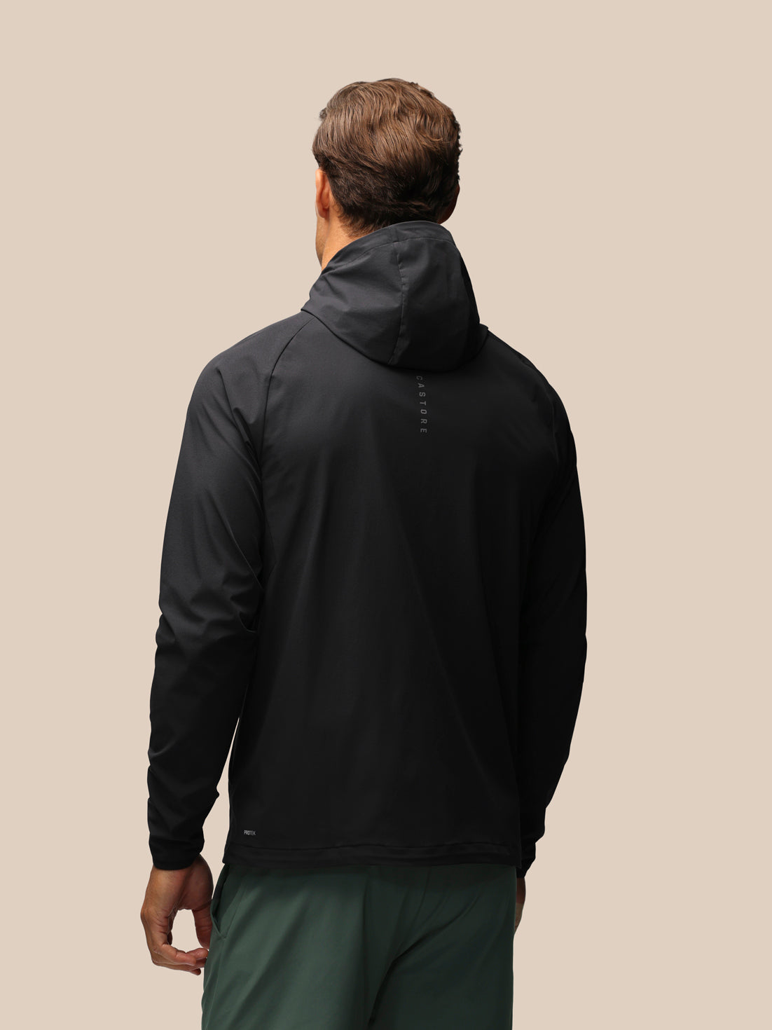 A man viewed from behind wearing a black hooded jacket and dark green pants against a solid beige background.
