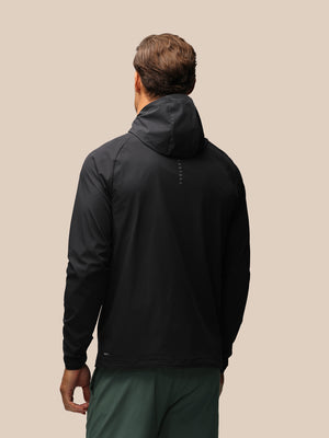 A man viewed from behind wearing a black hooded jacket and dark green pants against a solid beige background.