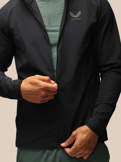 A close-up of a person wearing a black lightweight zip-up jacket with a subtle embroidered winged logo and the word 'CASTORE' on the left chest, over a muted green quarter-zip top and green bottoms, against a plain beige background.
