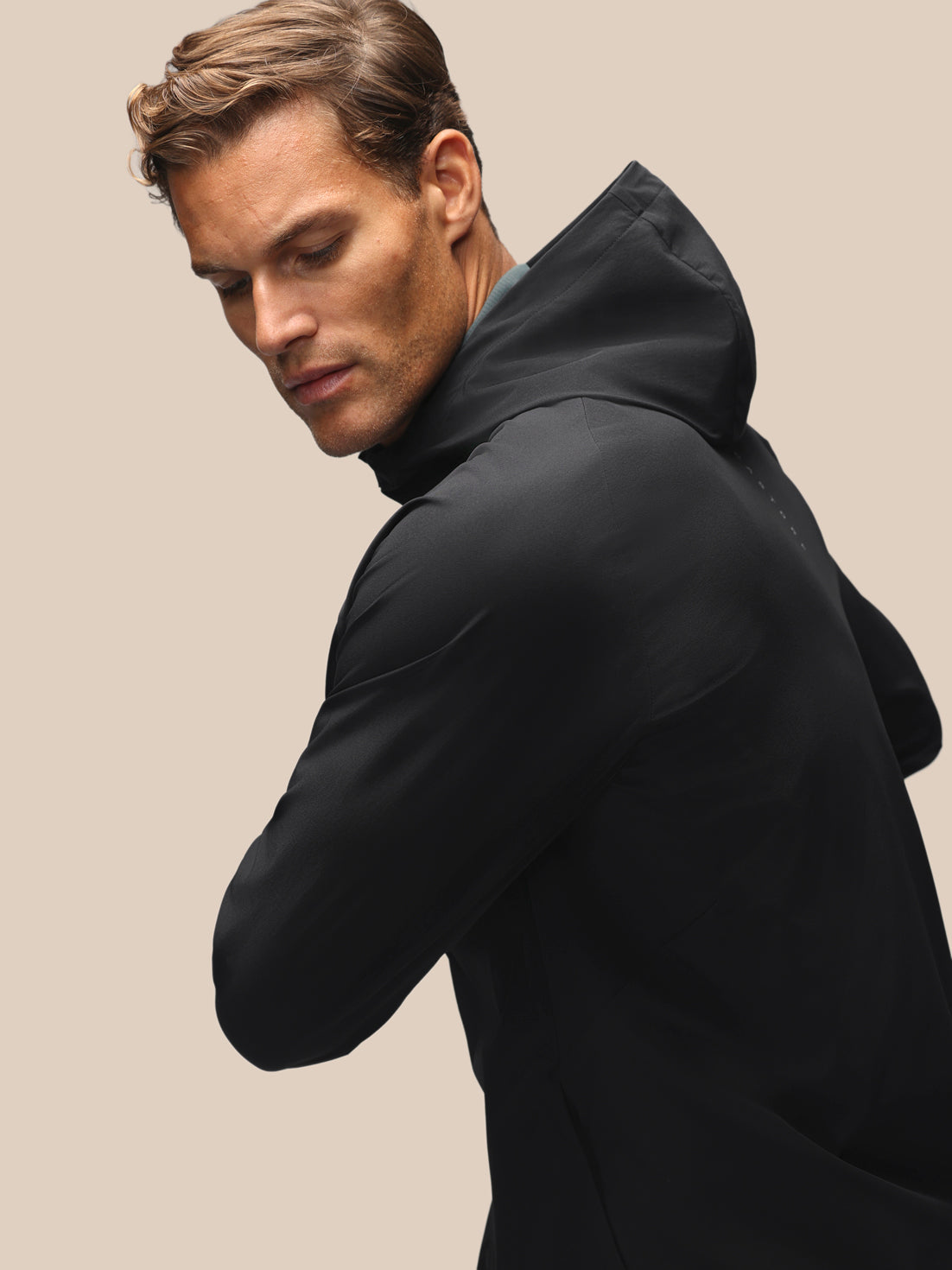 A man wearing a black hooded jacket, shown from a side-back angle against a solid beige background.