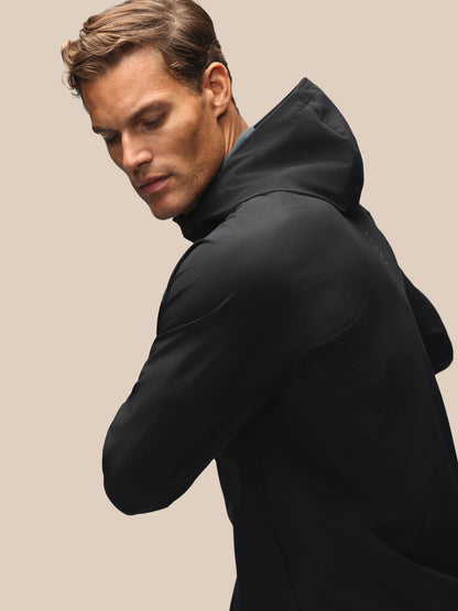 A man wearing a black hooded jacket, shown from a side-back angle against a solid beige background.
