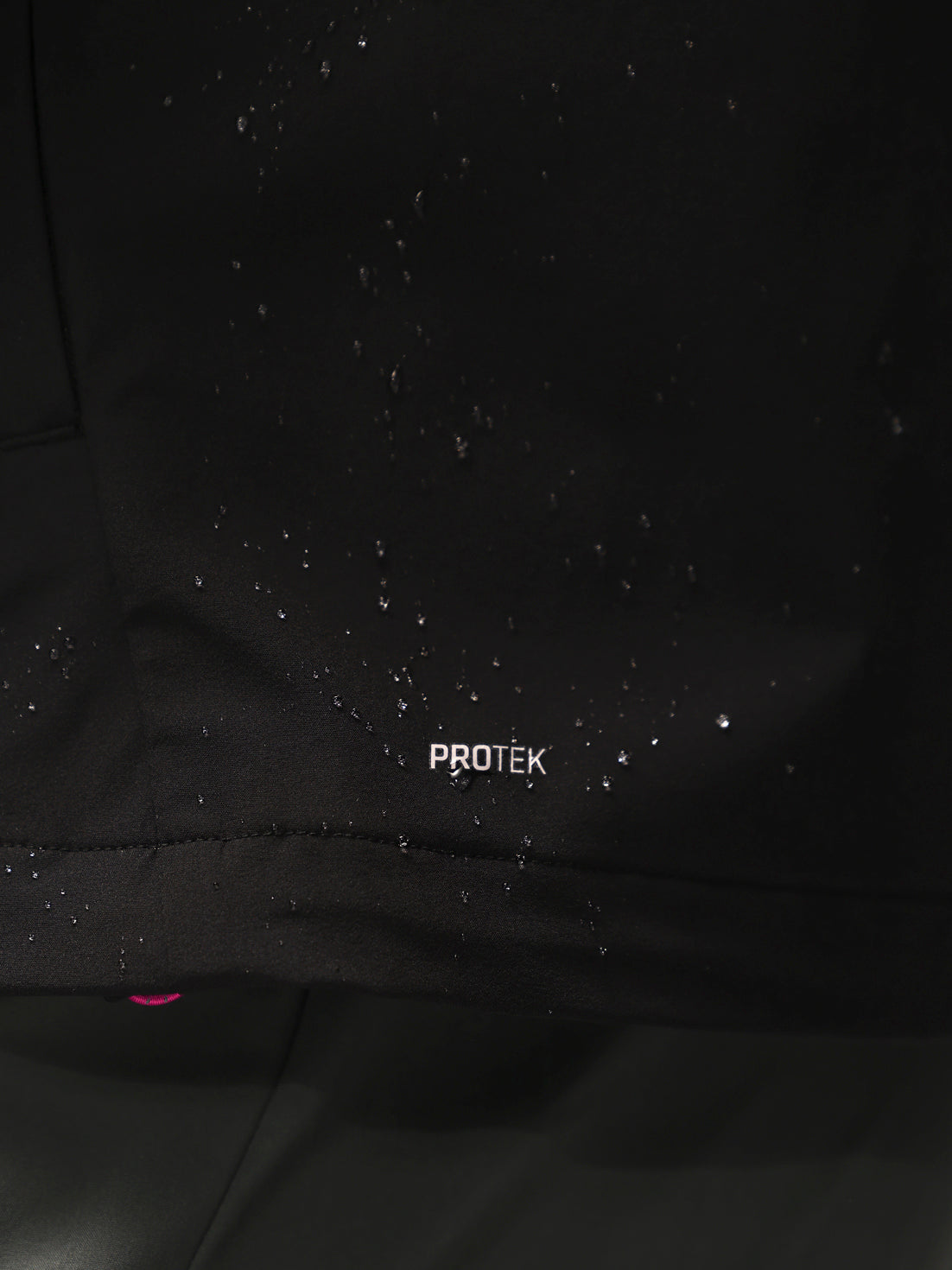 Close-up of a black waterproof jacket with water droplets beaded on the surface and the word 'PROTEK' printed in white near the bottom hem.