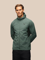 A man wearing a sage green hooded windbreaker jacket with black side panels and a small wing logo on the left chest, posed against a solid beige background.