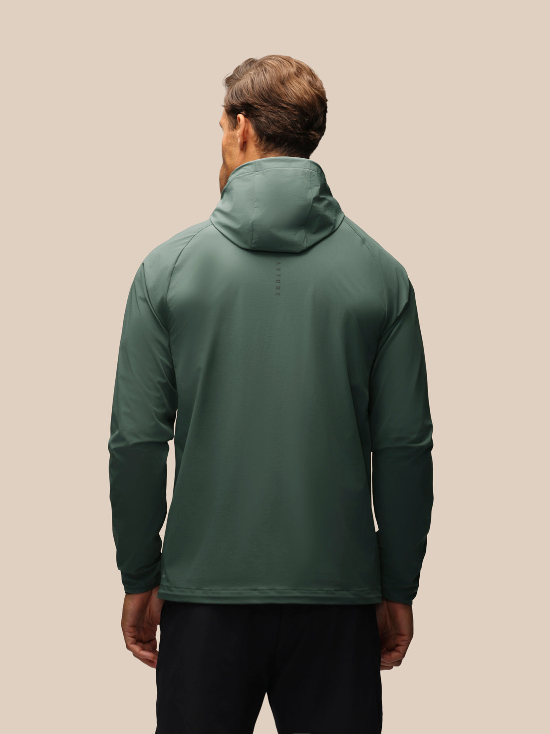 A man viewed from behind wearing a dark green hooded athletic jacket and black pants, standing against a solid beige background.