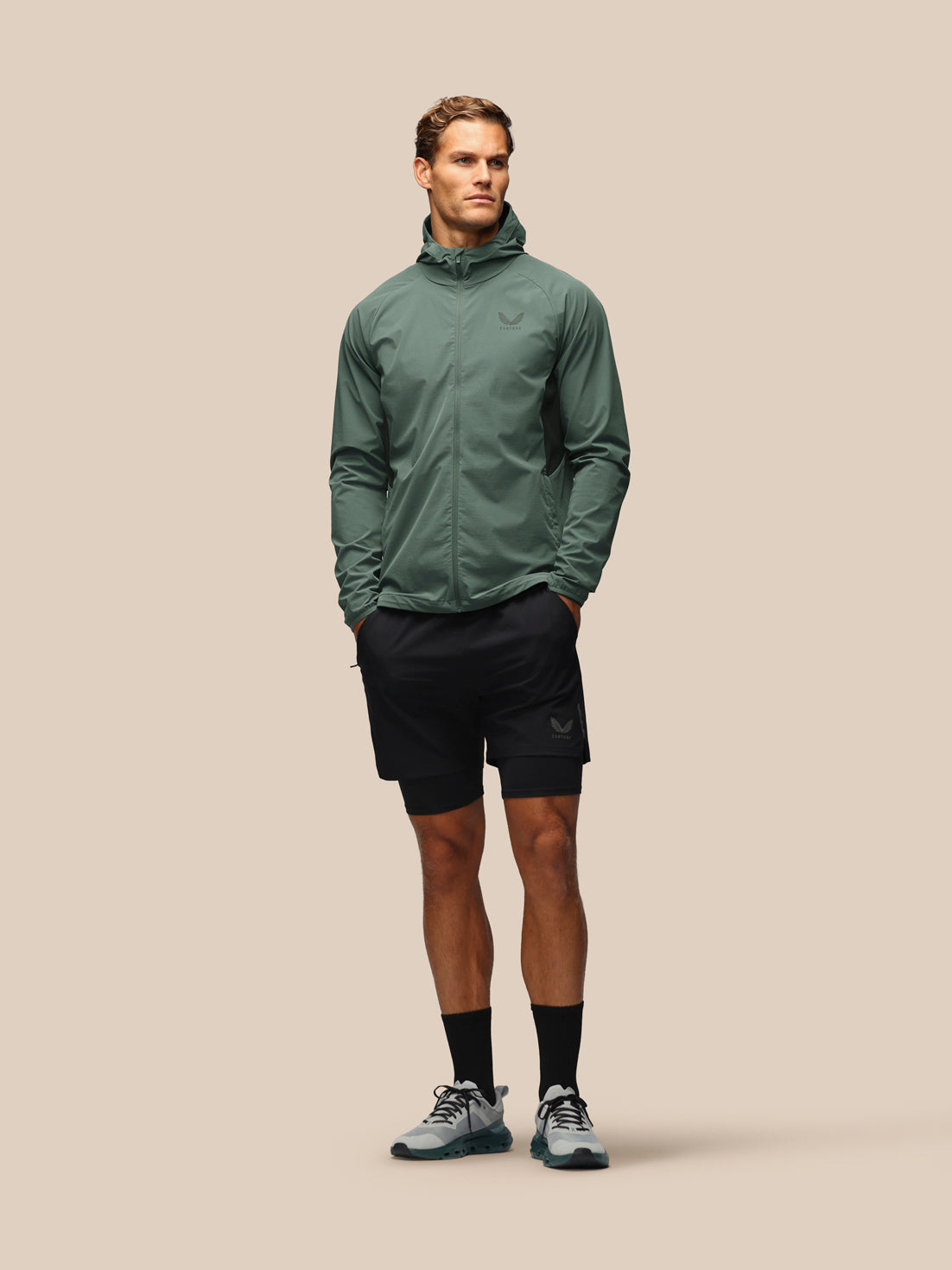 A male model stands against a solid beige background wearing a sage green lightweight zip-up hooded jacket, black athletic shorts, black socks, and gray running shoes.