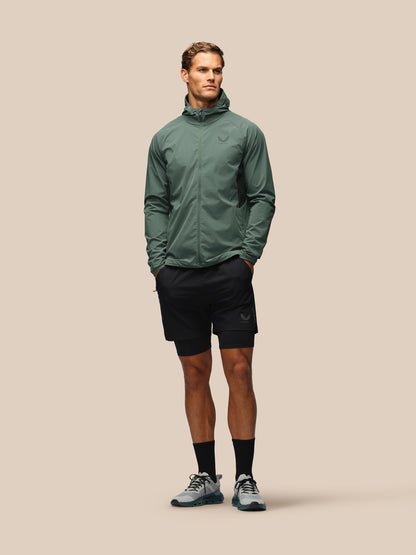 A male model stands against a solid beige background wearing a sage green lightweight zip-up hooded jacket, black athletic shorts, black socks, and gray running shoes.