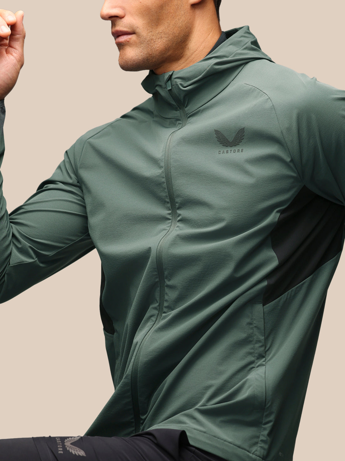 A man wearing a sage green athletic zip-up jacket with a black wing logo on the chest, photographed against a solid beige background.