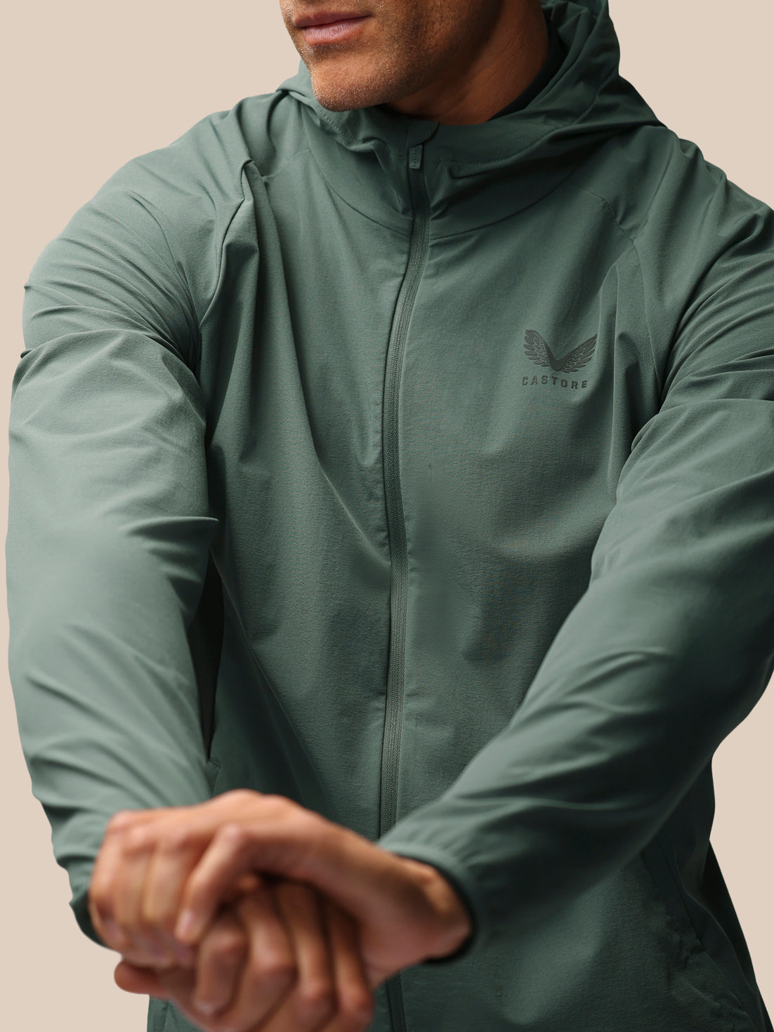 A man wearing a sage green hooded zip-up jacket with a subtle winged logo on the left chest, shown in a close-up shot against a solid beige background.