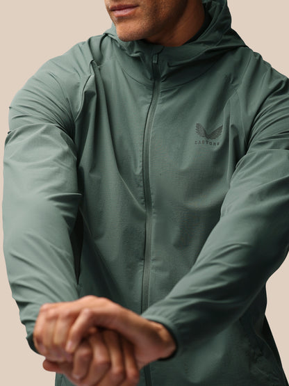 A man wearing a sage green hooded zip-up jacket with a subtle winged logo on the left chest, shown in a close-up shot against a solid beige background.
