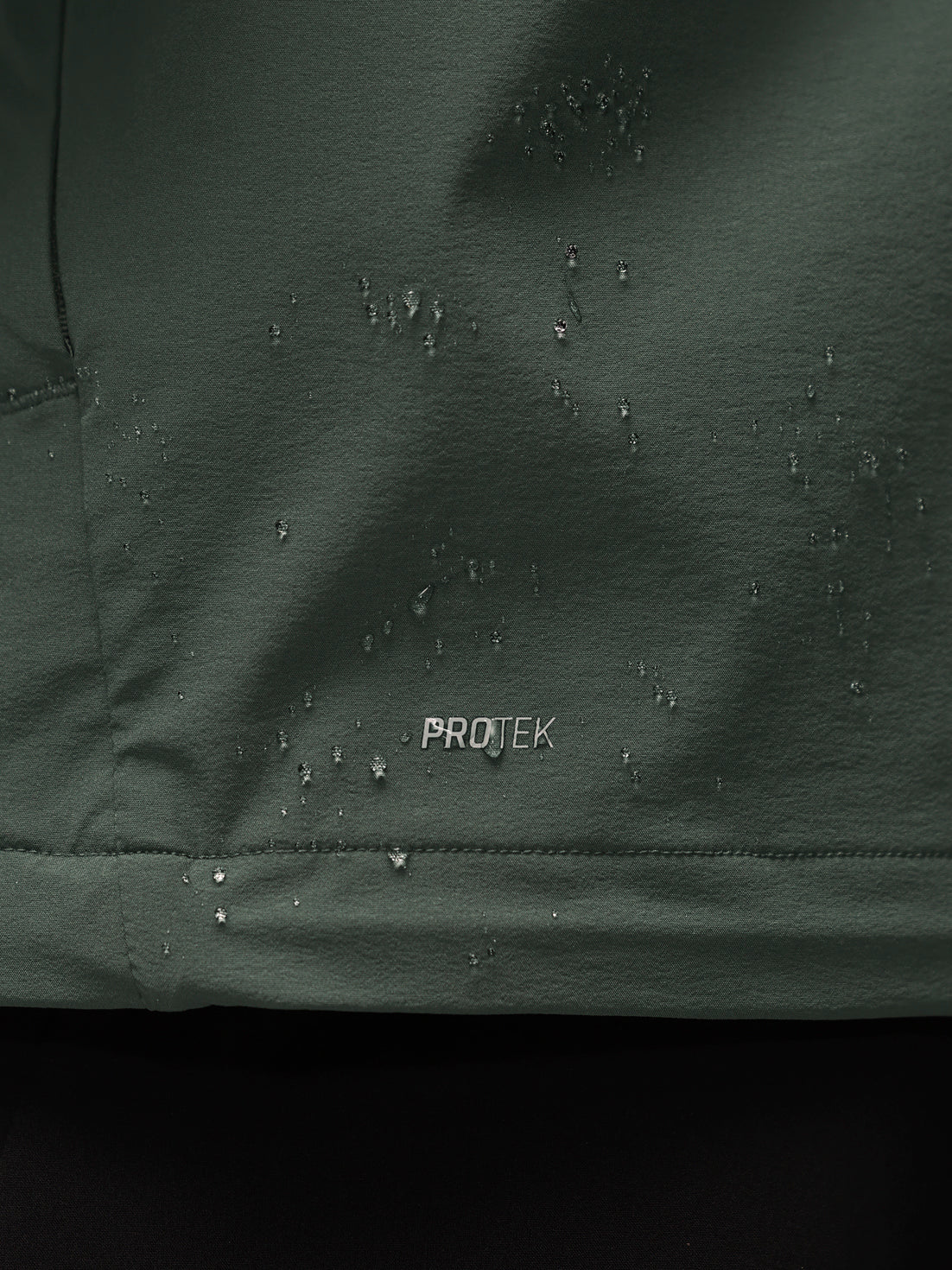 Close-up of a dark green waterproof fabric with water droplets beaded on the surface and the word PROTEK printed in white near the center.