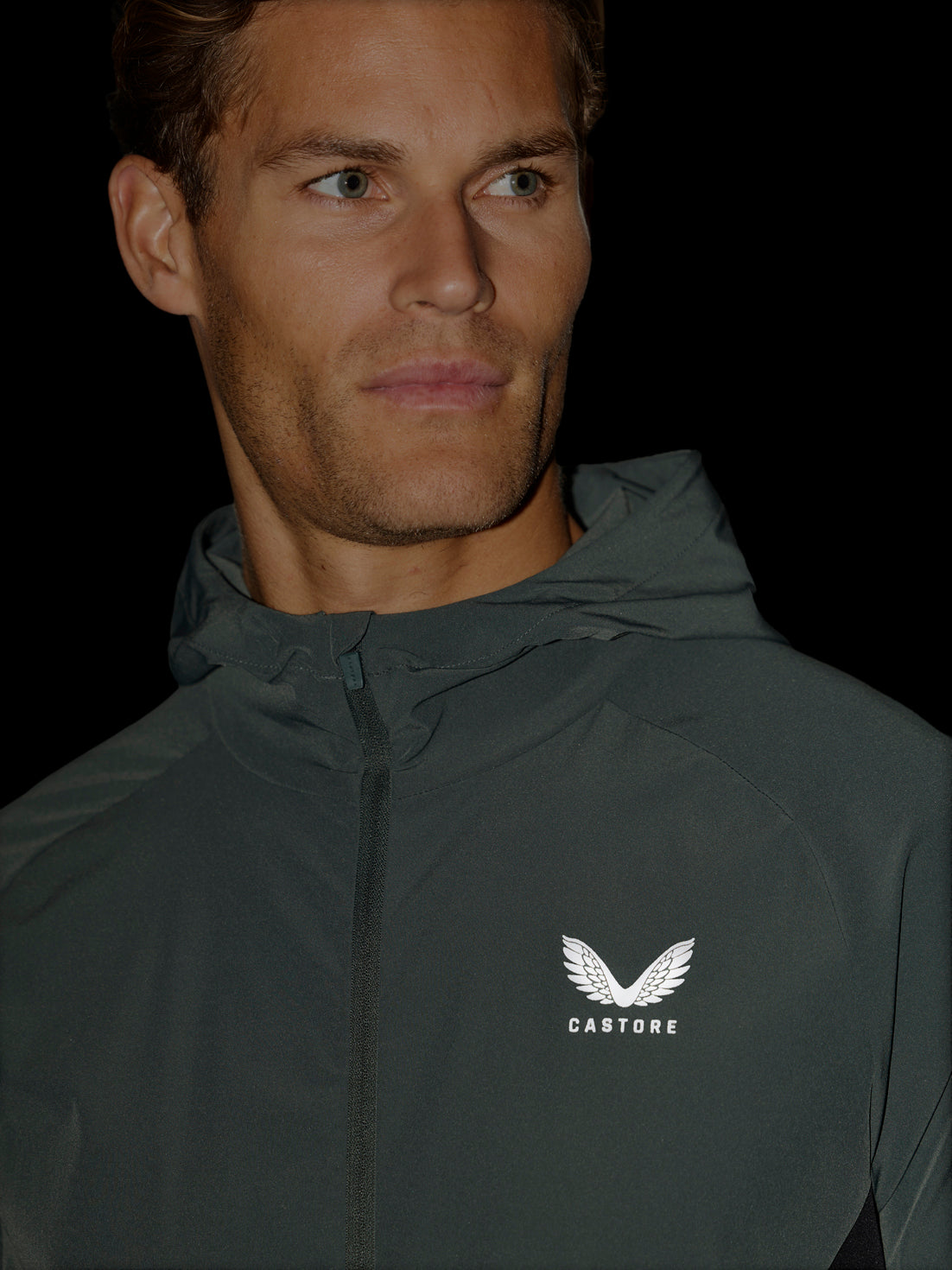 A close-up of a man wearing a dark gray hooded windbreaker jacket with a white wing logo and the brand name 'CASTORE' on the left chest, set against a solid black background.