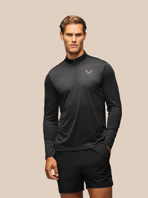A man modeling a black long-sleeve athletic quarter-zip pullover with a small wing logo on the left chest, paired with black shorts, against a solid beige background.