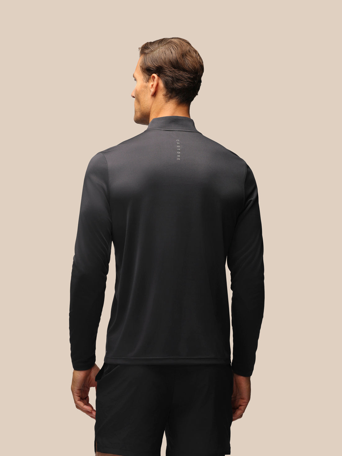 A man viewed from the back wearing a dark charcoal gray long-sleeve athletic quarter-zip top and black shorts against a solid beige background.