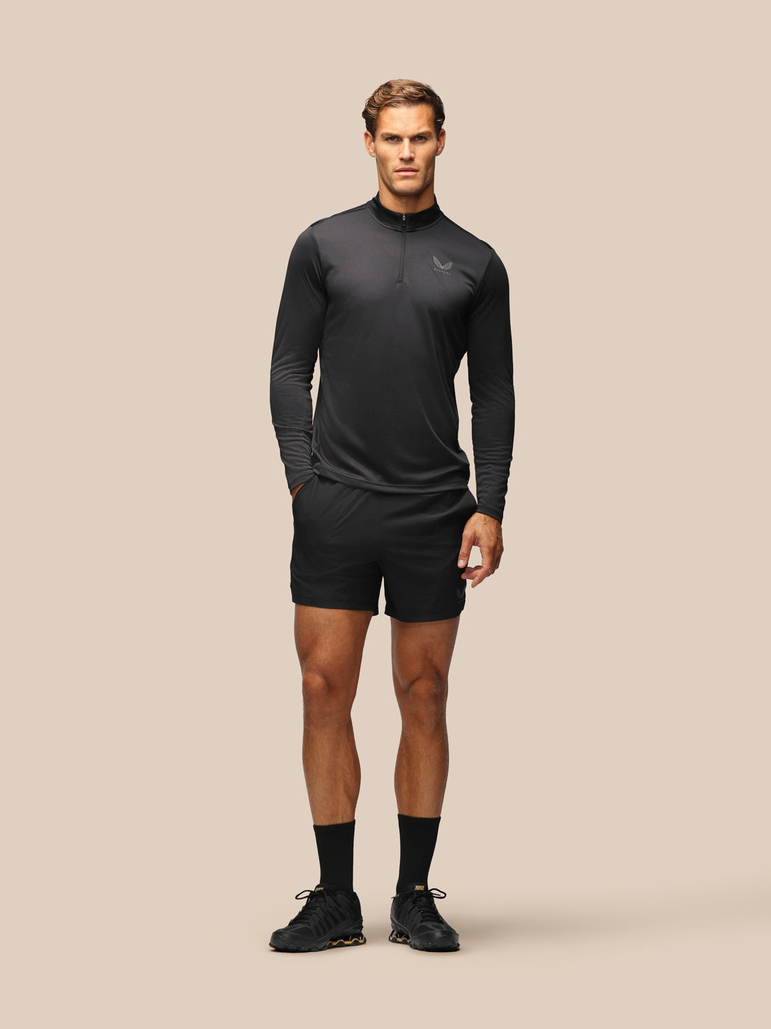 A male model stands against a solid beige studio background wearing a dark gray long-sleeve quarter-zip athletic top, black athletic shorts, black socks, and black running shoes.