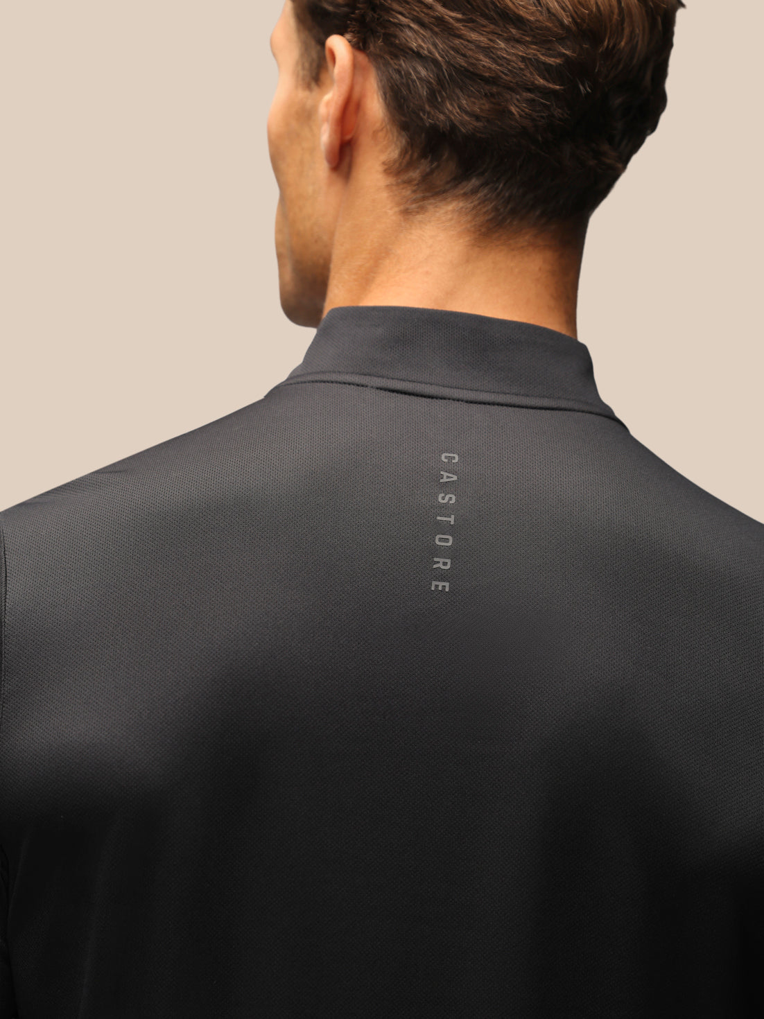 Close-up back view of a man wearing a dark charcoal gray mock neck shirt with the brand name 'CASTORE' printed vertically on the upper back, against a solid beige background.