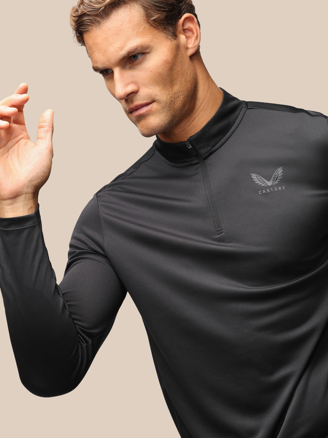 A man wearing a black long-sleeve athletic quarter-zip pullover with a subtle winged logo and the brand name 'CASTORE' on the left chest, set against a solid beige background.
