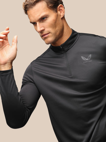 A man wearing a black long-sleeve athletic quarter-zip pullover with a subtle winged logo and the brand name 'CASTORE' on the left chest, set against a solid beige background.