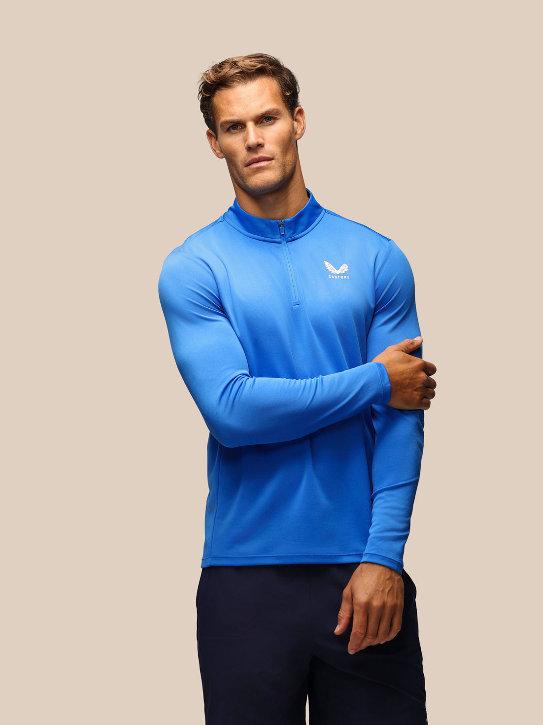 A man wearing a bright blue long-sleeve quarter-zip athletic top with a white wing logo on the left chest, paired with dark navy pants, set against a solid beige background.