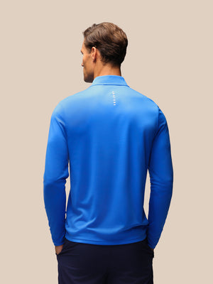 A man viewed from behind wearing a bright blue long-sleeve quarter-zip pullover with a small vertical white logo on the upper back, paired with dark navy pants, against a solid beige background.