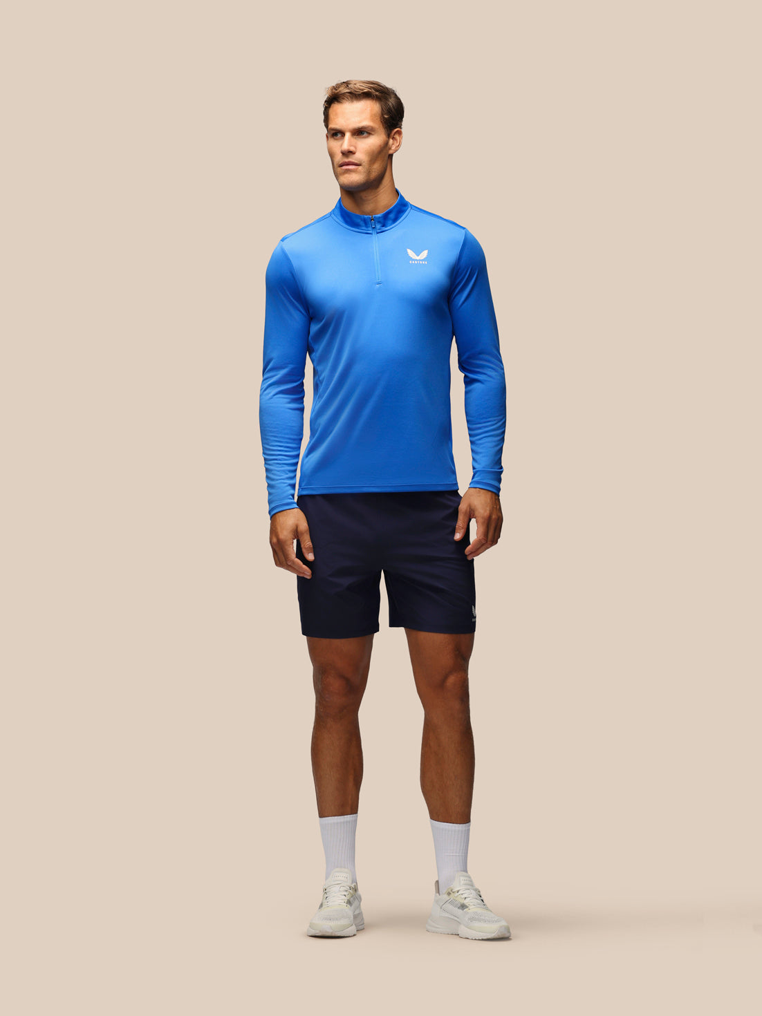 A male model stands against a solid beige background wearing a bright blue long-sleeve athletic quarter-zip top, dark navy athletic shorts, white socks, and white sneakers.