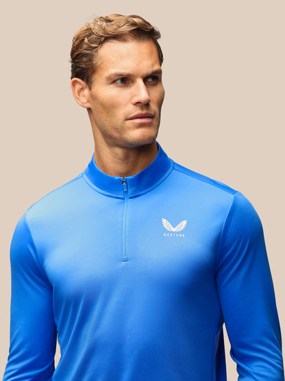 A man wearing a bright blue long-sleeve athletic quarter-zip pullover with a white wing logo and the brand name 'CASTORE' on the left chest, set against a solid beige background.