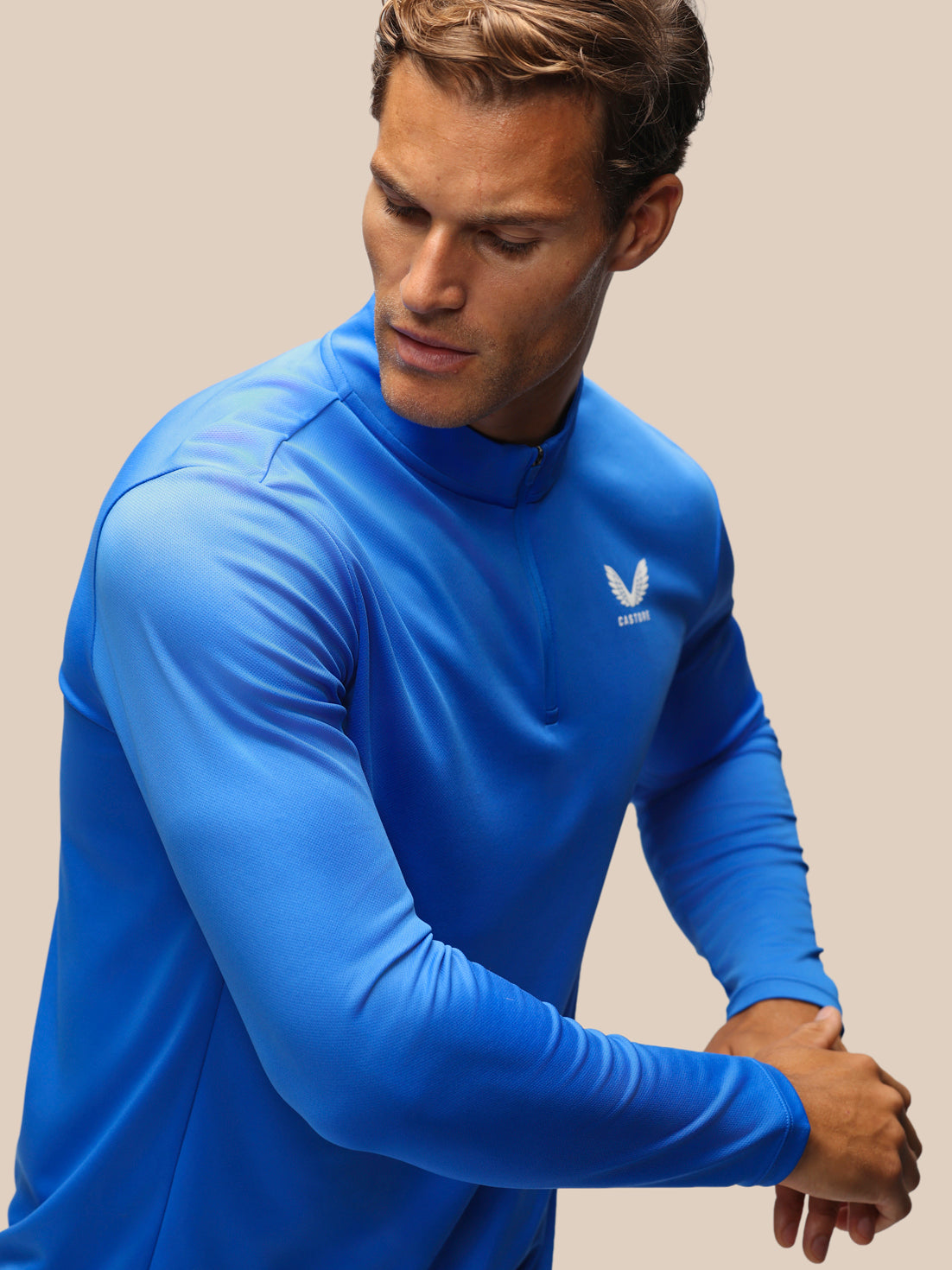 A man wearing a bright blue long-sleeve athletic quarter-zip pullover with a small white wing logo on the left chest, posed against a solid beige background.