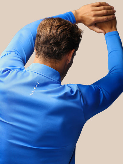A man wearing a bright blue long-sleeve athletic shirt, shown from behind with his arms raised, on a plain beige background. The brand name 'CASTORE' is printed vertically on the back of the shirt.