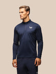 A man wearing a navy blue long-sleeve athletic quarter-zip pullover with a white logo on the left chest, paired with matching navy pants, set against a solid beige background.