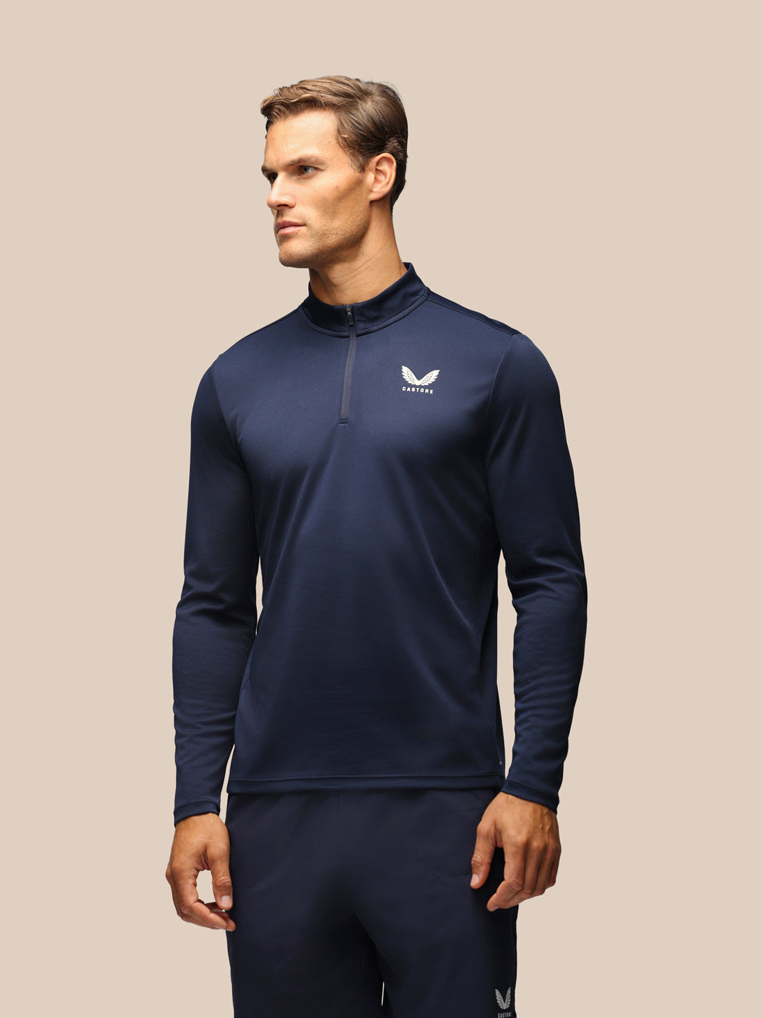 A man wearing a navy blue long-sleeve athletic quarter-zip pullover with a white logo on the left chest, paired with matching navy pants, set against a solid beige background.