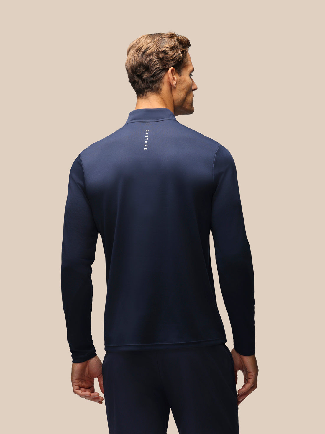 A man shown from the back wearing a navy blue long-sleeve athletic quarter-zip top and matching navy pants against a solid beige background.
