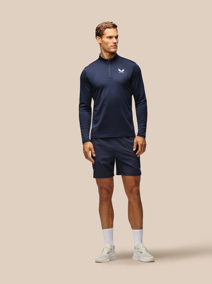 A man modeling a navy blue athletic half-zip pullover and matching shorts set, featuring a small white logo on the chest, against a solid beige background.