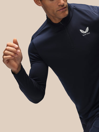 A man wearing a navy blue long-sleeve athletic quarter-zip pullover with a white winged logo and the word 'CASTORE' on the left chest, set against a solid beige background.