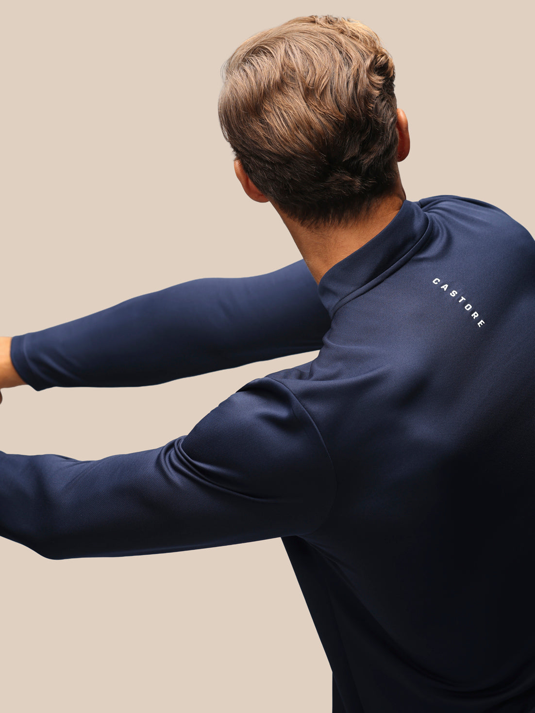 A man wearing a navy blue long-sleeve athletic top, shown from the back and side against a solid beige background. The brand name 'CASTORE' is printed in white on the upper shoulder.