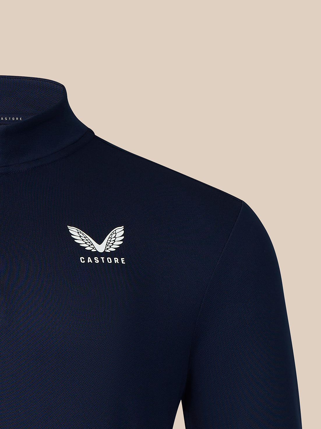 Close-up of a navy blue mock neck sweater with a white embroidered winged logo and the brand name 'CASTORE' on the left chest, set against a solid beige background.